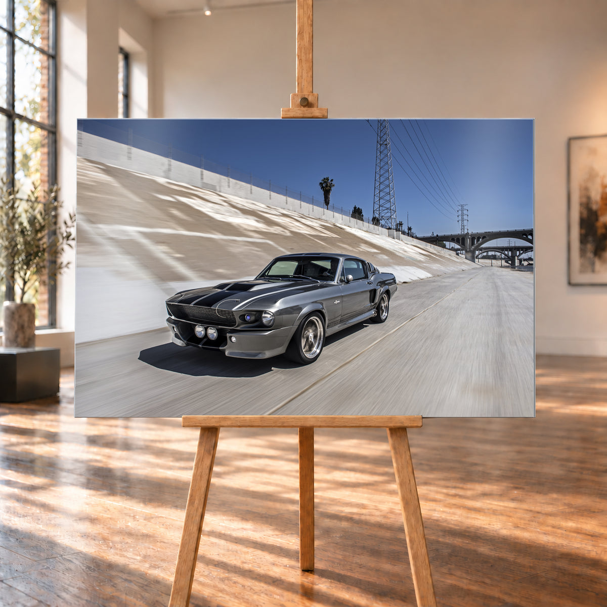 Metallisch grauer Ford Mustang Shelby GT500 mit schwarzen Doppelstreifen in niedriger Front-Dreiviertelansicht in einem hellen Betonkanal. Fahrbahn und Kanalwand sind bewegungsunscharf; Strommasten, Leitungen, Palmen und Bogenbrücken stehen unter blauem Himmel; Visualisierung als Alu gebürstet-Wandbild, auf einer Holzstaffelei in einem hellen Galerieraum.