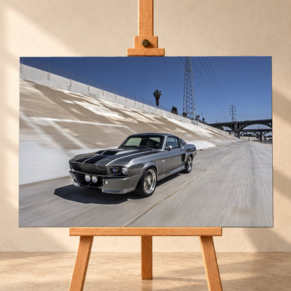 Metallisch grauer Ford Mustang Shelby GT500 mit schwarzen Doppelstreifen in niedriger Front-Dreiviertelansicht in einem hellen Betonkanal. Fahrbahn und Kanalwand sind bewegungsunscharf; Strommasten, Leitungen, Palmen und Bogenbrücken stehen unter blauem Himmel; Visualisierung als Leinwand-Wandbild, frontal auf einer Holzstaffelei.