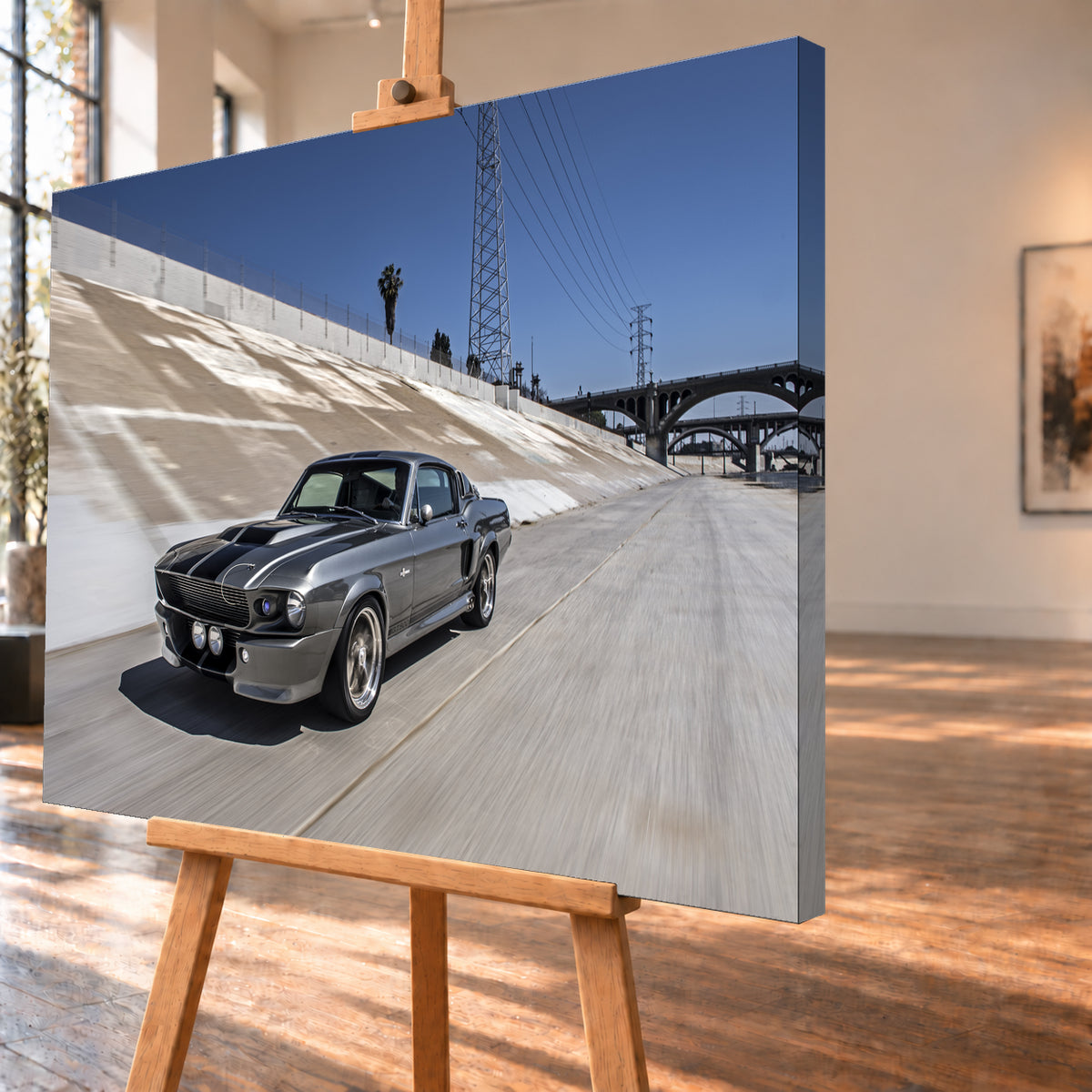 Metallisch grauer Ford Mustang Shelby GT500 mit schwarzen Doppelstreifen in niedriger Front-Dreiviertelansicht in einem hellen Betonkanal. Fahrbahn und Kanalwand sind bewegungsunscharf; Strommasten, Leitungen, Palmen und Bogenbrücken stehen unter blauem Himmel; Visualisierung als Leinwand-Wandbild, schräg von der Seite auf einer Holzstaffelei.