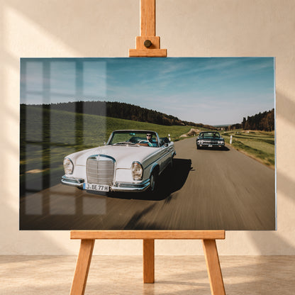 Frontale Fahraufnahme eines weißen 1965 Mercedes 300 SE W112 Cabriolets im Vordergrund und eines dunklen 1964 Lincoln Continental Cabriolets dahinter auf einer Landstraße. Grüne Felder, Waldlinien, blauer Himmel und Bewegungsunschärfe umgeben die beiden offenen Fahrzeuge im Sonnenlicht; Visualisierung als Acrylglas-Wandbild, frontal auf einer Holzstaffelei.