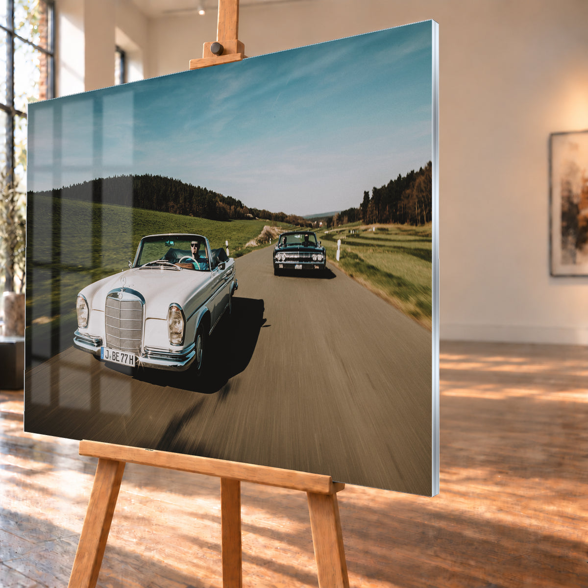 Frontale Fahraufnahme eines weißen 1965 Mercedes 300 SE W112 Cabriolets im Vordergrund und eines dunklen 1964 Lincoln Continental Cabriolets dahinter auf einer Landstraße. Grüne Felder, Waldlinien, blauer Himmel und Bewegungsunschärfe umgeben die beiden offenen Fahrzeuge im Sonnenlicht; Visualisierung als Acrylglas-Wandbild, schräg von der Seite auf einer Holzstaffelei.