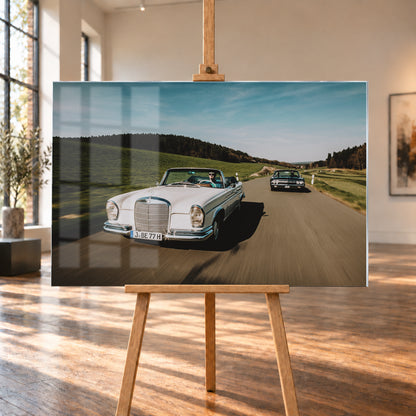 Frontale Fahraufnahme eines weißen 1965 Mercedes 300 SE W112 Cabriolets im Vordergrund und eines dunklen 1964 Lincoln Continental Cabriolets dahinter auf einer Landstraße. Grüne Felder, Waldlinien, blauer Himmel und Bewegungsunschärfe umgeben die beiden offenen Fahrzeuge im Sonnenlicht; Visualisierung als Acrylglas-Wandbild, auf einer Holzstaffelei in einem hellen Galerieraum.