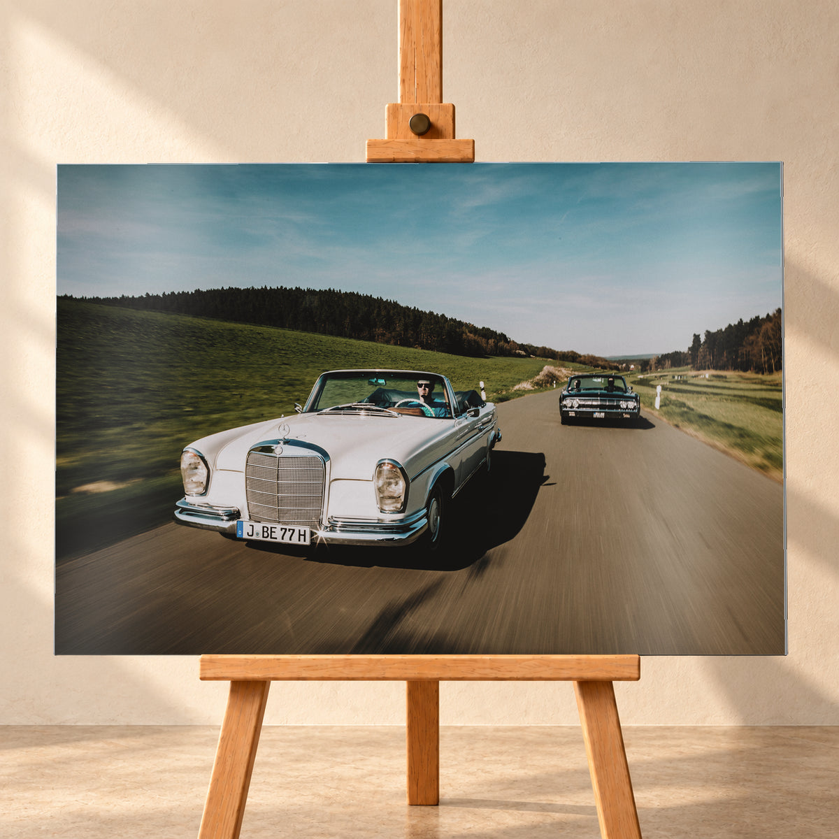 Frontale Fahraufnahme eines weißen 1965 Mercedes 300 SE W112 Cabriolets im Vordergrund und eines dunklen 1964 Lincoln Continental Cabriolets dahinter auf einer Landstraße. Grüne Felder, Waldlinien, blauer Himmel und Bewegungsunschärfe umgeben die beiden offenen Fahrzeuge im Sonnenlicht; Visualisierung als Alu gebürstet-Wandbild, frontal auf einer Holzstaffelei.
