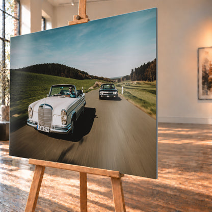 Frontale Fahraufnahme eines weißen 1965 Mercedes 300 SE W112 Cabriolets im Vordergrund und eines dunklen 1964 Lincoln Continental Cabriolets dahinter auf einer Landstraße. Grüne Felder, Waldlinien, blauer Himmel und Bewegungsunschärfe umgeben die beiden offenen Fahrzeuge im Sonnenlicht; Visualisierung als Alu gebürstet-Wandbild, schräg von der Seite auf einer Holzstaffelei.