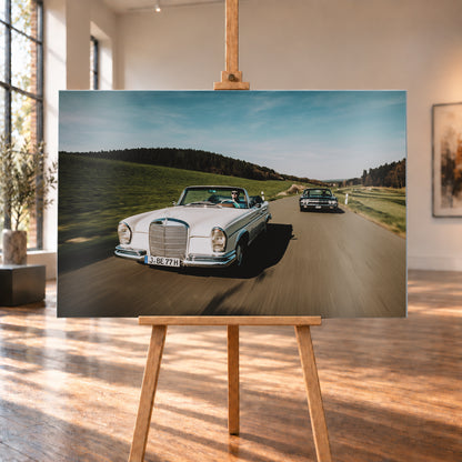 Frontale Fahraufnahme eines weißen 1965 Mercedes 300 SE W112 Cabriolets im Vordergrund und eines dunklen 1964 Lincoln Continental Cabriolets dahinter auf einer Landstraße. Grüne Felder, Waldlinien, blauer Himmel und Bewegungsunschärfe umgeben die beiden offenen Fahrzeuge im Sonnenlicht; Visualisierung als Alu gebürstet-Wandbild, auf einer Holzstaffelei in einem hellen Galerieraum.
