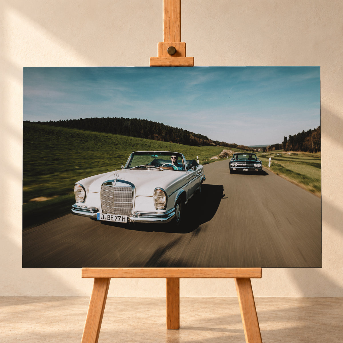 Frontale Fahraufnahme eines weißen 1965 Mercedes 300 SE W112 Cabriolets im Vordergrund und eines dunklen 1964 Lincoln Continental Cabriolets dahinter auf einer Landstraße. Grüne Felder, Waldlinien, blauer Himmel und Bewegungsunschärfe umgeben die beiden offenen Fahrzeuge im Sonnenlicht; Visualisierung als Leinwand-Wandbild, frontal auf einer Holzstaffelei.