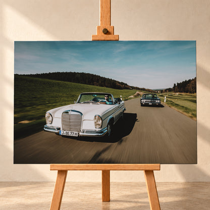 Frontale Fahraufnahme eines weißen 1965 Mercedes 300 SE W112 Cabriolets im Vordergrund und eines dunklen 1964 Lincoln Continental Cabriolets dahinter auf einer Landstraße. Grüne Felder, Waldlinien, blauer Himmel und Bewegungsunschärfe umgeben die beiden offenen Fahrzeuge im Sonnenlicht; Visualisierung als Leinwand-Wandbild, frontal auf einer Holzstaffelei.