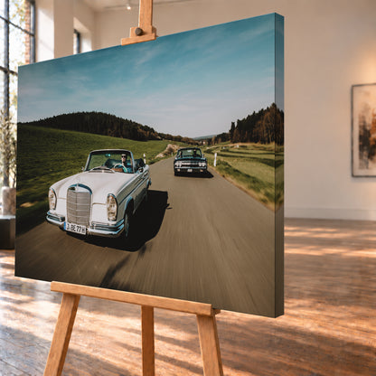 Frontale Fahraufnahme eines weißen 1965 Mercedes 300 SE W112 Cabriolets im Vordergrund und eines dunklen 1964 Lincoln Continental Cabriolets dahinter auf einer Landstraße. Grüne Felder, Waldlinien, blauer Himmel und Bewegungsunschärfe umgeben die beiden offenen Fahrzeuge im Sonnenlicht; Visualisierung als Leinwand-Wandbild, schräg von der Seite auf einer Holzstaffelei.