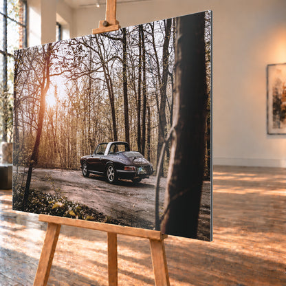 Dunkler 1967 Porsche 911 S Softwindow in seitlicher Heckansicht auf einer unbefestigten Fläche im Wald. Die offene Dachpartie und die helle Bügelzone sind sichtbar, während tiefes Sonnenlicht links durch hohe, spärlich belaubte Bäume fällt; Visualisierung als Alu gebürstet-Wandbild, schräg von der Seite auf einer Holzstaffelei.