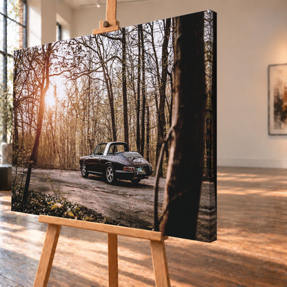 Dunkler 1967 Porsche 911 S Softwindow in seitlicher Heckansicht auf einer unbefestigten Fläche im Wald. Die offene Dachpartie und die helle Bügelzone sind sichtbar, während tiefes Sonnenlicht links durch hohe, spärlich belaubte Bäume fällt; Visualisierung als Leinwand-Wandbild, schräg von der Seite auf einer Holzstaffelei.