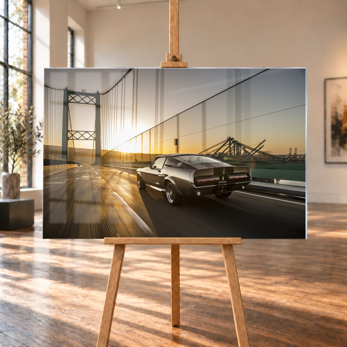 Dunkelgrauer Ford Mustang Fastback in Heck-Dreiviertelansicht auf einer Hängebrücke; goldenes Gegenlicht spiegelt sich auf der Fahrbahn, rechts sind Wasser und Hafenkräne zu sehen; Visualisierung als Acrylglas-Wandbild, auf einer Holzstaffelei in einem hellen Galerieraum.