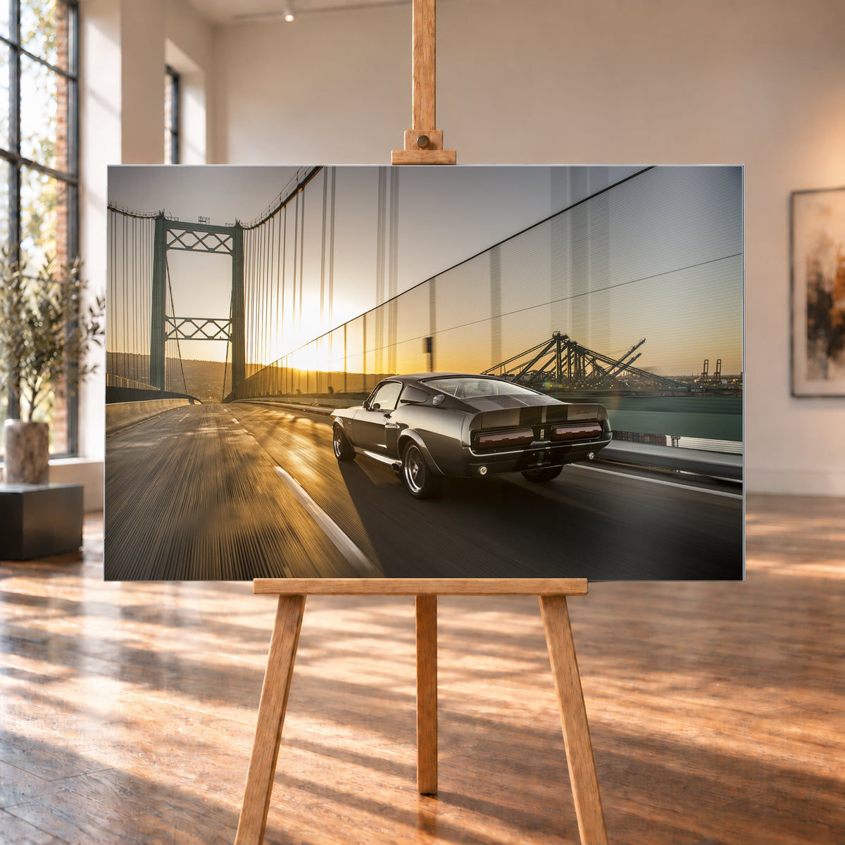 Dunkelgrauer Ford Mustang Fastback in Heck-Dreiviertelansicht auf einer Hängebrücke; goldenes Gegenlicht spiegelt sich auf der Fahrbahn, rechts sind Wasser und Hafenkräne zu sehen; Visualisierung als Alu gebürstet-Wandbild, auf einer Holzstaffelei in einem hellen Galerieraum.