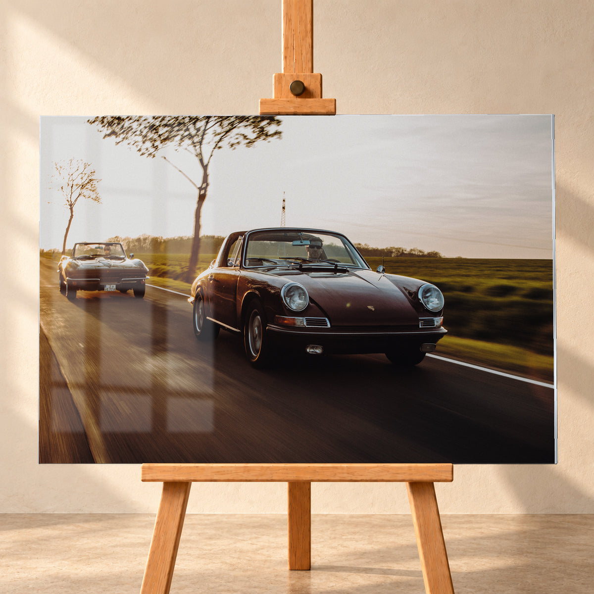 Ein dunkelrot wirkender Porsche 911 S Softwindow fährt rechts im Vordergrund, dahinter links eine dunkle Chevrolet Corvette C2; beide mit offenem Dach und sichtbaren Fahrern auf einer Landstraße bei tiefem, warmem Gegenlicht; Visualisierung als Acrylglas-Wandbild, frontal auf einer Holzstaffelei.
