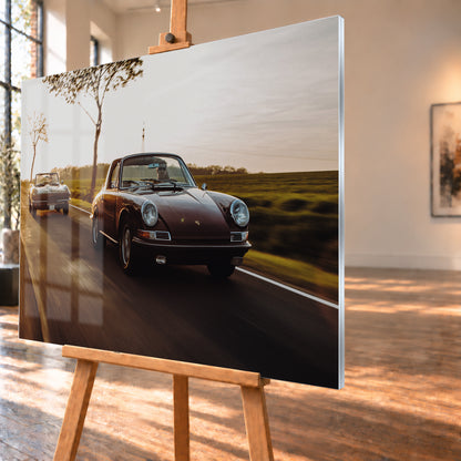 Ein dunkelrot wirkender Porsche 911 S Softwindow fährt rechts im Vordergrund, dahinter links eine dunkle Chevrolet Corvette C2; beide mit offenem Dach und sichtbaren Fahrern auf einer Landstraße bei tiefem, warmem Gegenlicht; Visualisierung als Acrylglas-Wandbild, schräg von der Seite auf einer Holzstaffelei.