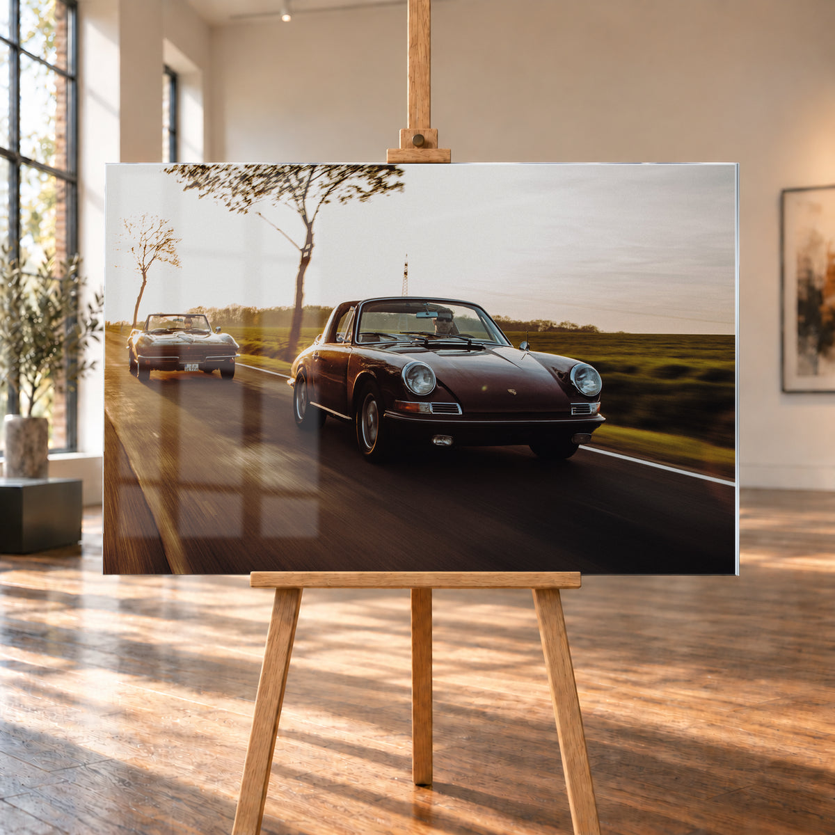 Ein dunkelrot wirkender Porsche 911 S Softwindow fährt rechts im Vordergrund, dahinter links eine dunkle Chevrolet Corvette C2; beide mit offenem Dach und sichtbaren Fahrern auf einer Landstraße bei tiefem, warmem Gegenlicht; Visualisierung als Acrylglas-Wandbild, auf einer Holzstaffelei in einem hellen Galerieraum.