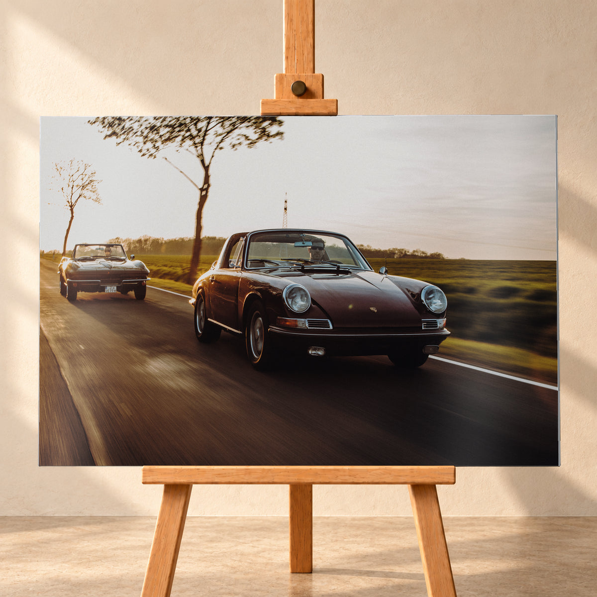 Ein dunkelrot wirkender Porsche 911 S Softwindow fährt rechts im Vordergrund, dahinter links eine dunkle Chevrolet Corvette C2; beide mit offenem Dach und sichtbaren Fahrern auf einer Landstraße bei tiefem, warmem Gegenlicht; Visualisierung als Alu gebürstet-Wandbild, frontal auf einer Holzstaffelei.