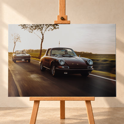 Ein dunkelrot wirkender Porsche 911 S Softwindow fährt rechts im Vordergrund, dahinter links eine dunkle Chevrolet Corvette C2; beide mit offenem Dach und sichtbaren Fahrern auf einer Landstraße bei tiefem, warmem Gegenlicht; Visualisierung als Alu gebürstet-Wandbild, frontal auf einer Holzstaffelei.