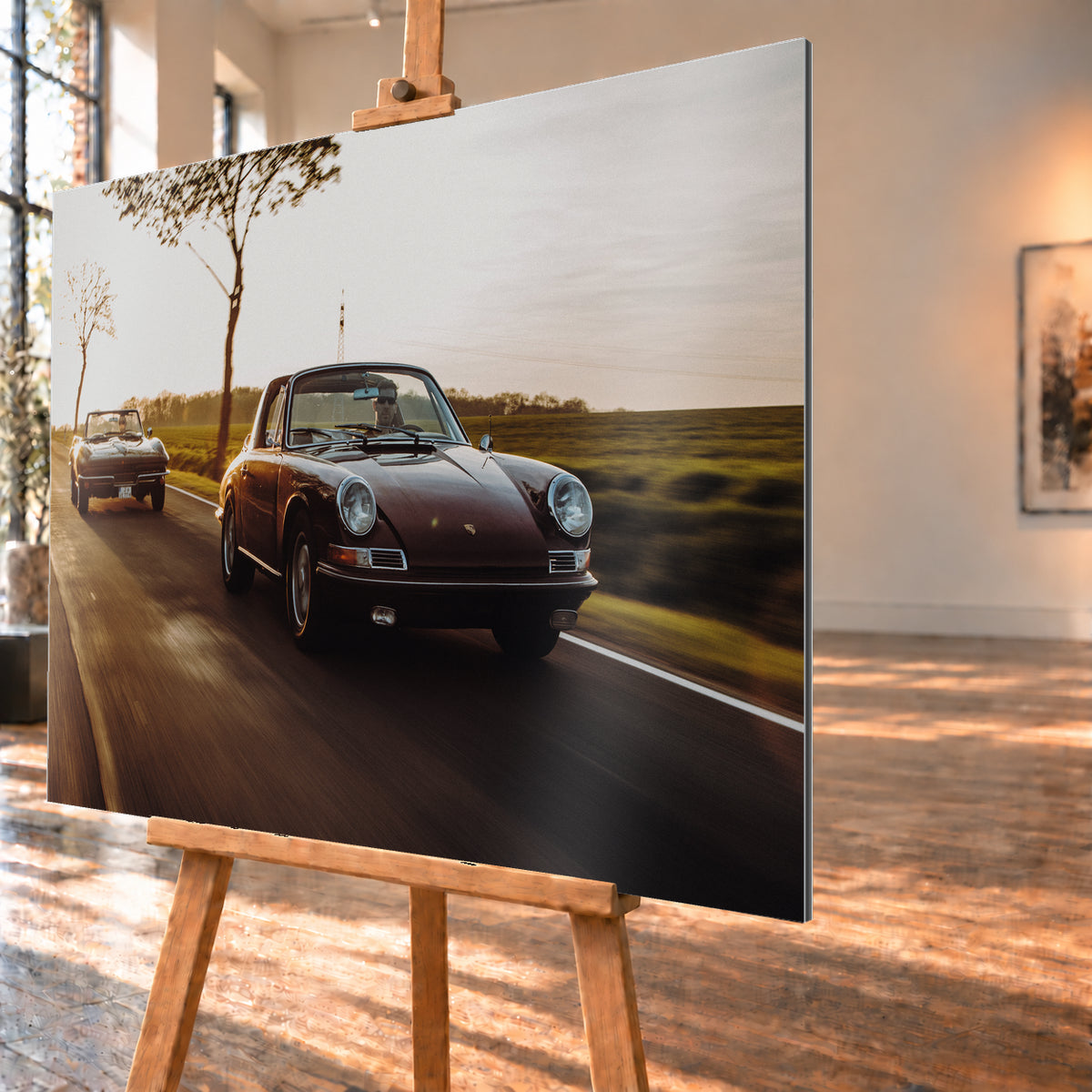 Ein dunkelrot wirkender Porsche 911 S Softwindow fährt rechts im Vordergrund, dahinter links eine dunkle Chevrolet Corvette C2; beide mit offenem Dach und sichtbaren Fahrern auf einer Landstraße bei tiefem, warmem Gegenlicht; Visualisierung als Alu gebürstet-Wandbild, schräg von der Seite auf einer Holzstaffelei.