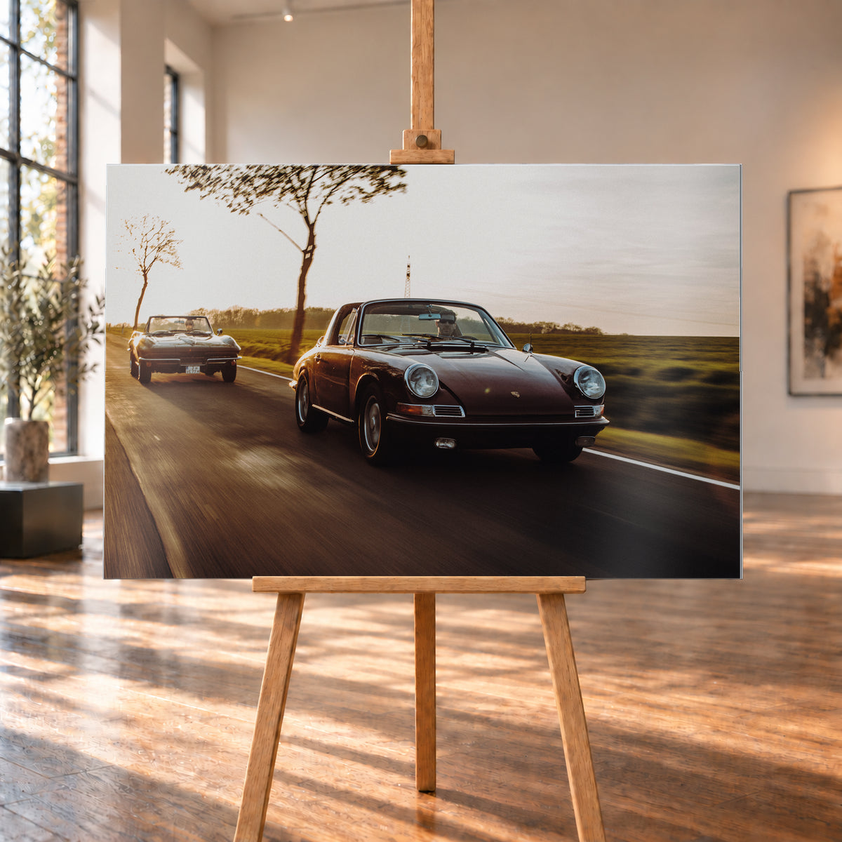 Ein dunkelrot wirkender Porsche 911 S Softwindow fährt rechts im Vordergrund, dahinter links eine dunkle Chevrolet Corvette C2; beide mit offenem Dach und sichtbaren Fahrern auf einer Landstraße bei tiefem, warmem Gegenlicht; Visualisierung als Alu gebürstet-Wandbild, auf einer Holzstaffelei in einem hellen Galerieraum.