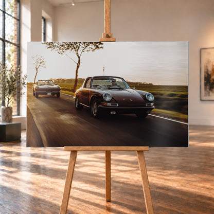 Ein dunkelrot wirkender Porsche 911 S Softwindow fährt rechts im Vordergrund, dahinter links eine dunkle Chevrolet Corvette C2; beide mit offenem Dach und sichtbaren Fahrern auf einer Landstraße bei tiefem, warmem Gegenlicht; Visualisierung als Alu gebürstet-Wandbild, auf einer Holzstaffelei in einem hellen Galerieraum.