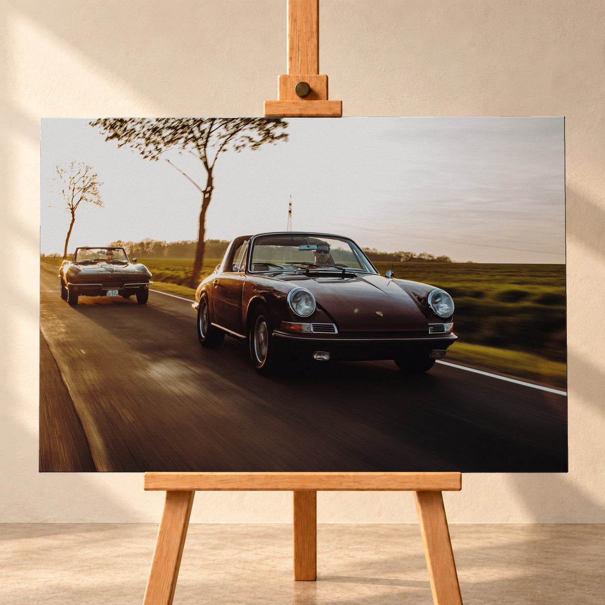Ein dunkelrot wirkender Porsche 911 S Softwindow fährt rechts im Vordergrund, dahinter links eine dunkle Chevrolet Corvette C2; beide mit offenem Dach und sichtbaren Fahrern auf einer Landstraße bei tiefem, warmem Gegenlicht; Visualisierung als Leinwand-Wandbild, frontal auf einer Holzstaffelei.