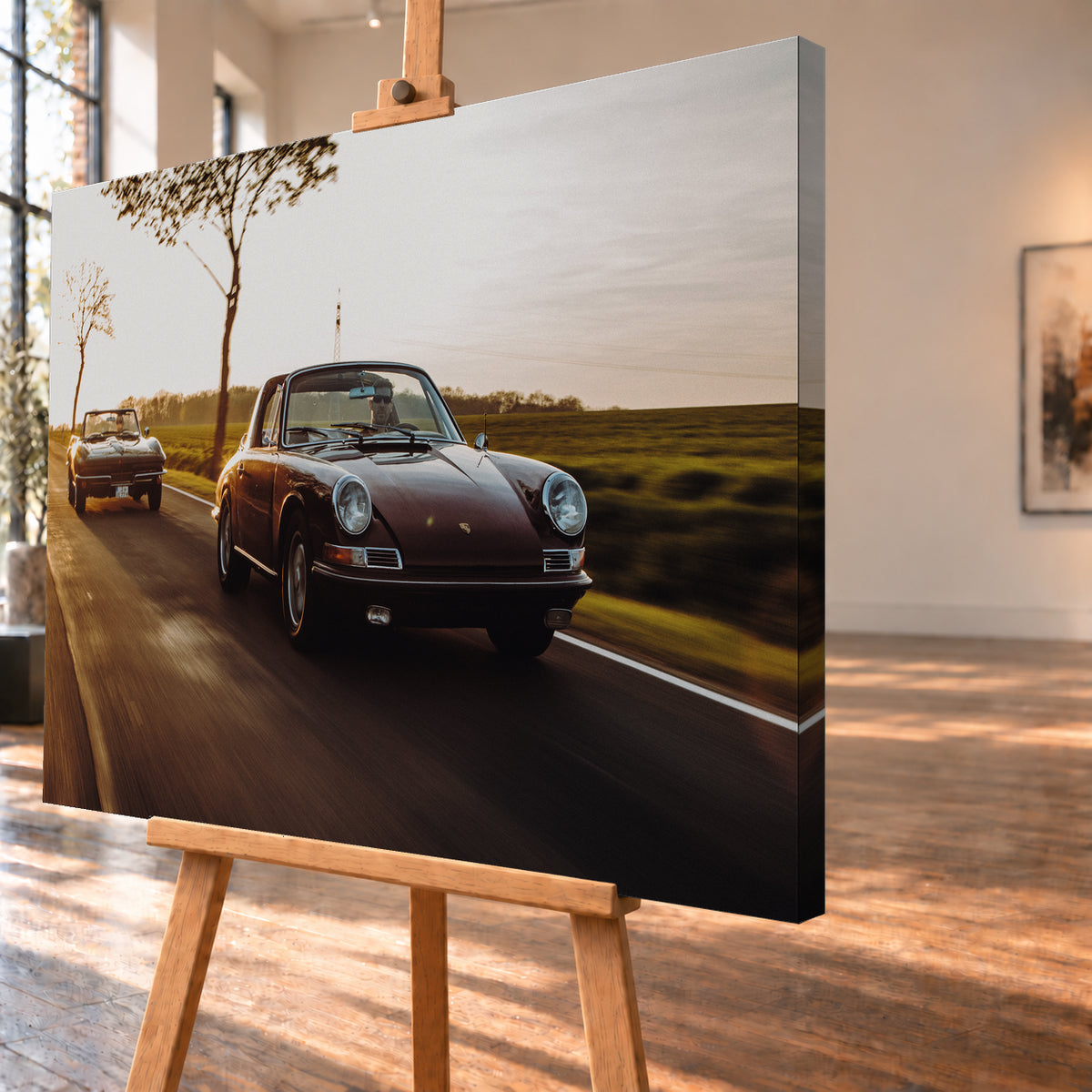Ein dunkelrot wirkender Porsche 911 S Softwindow fährt rechts im Vordergrund, dahinter links eine dunkle Chevrolet Corvette C2; beide mit offenem Dach und sichtbaren Fahrern auf einer Landstraße bei tiefem, warmem Gegenlicht; Visualisierung als Leinwand-Wandbild, schräg von der Seite auf einer Holzstaffelei.