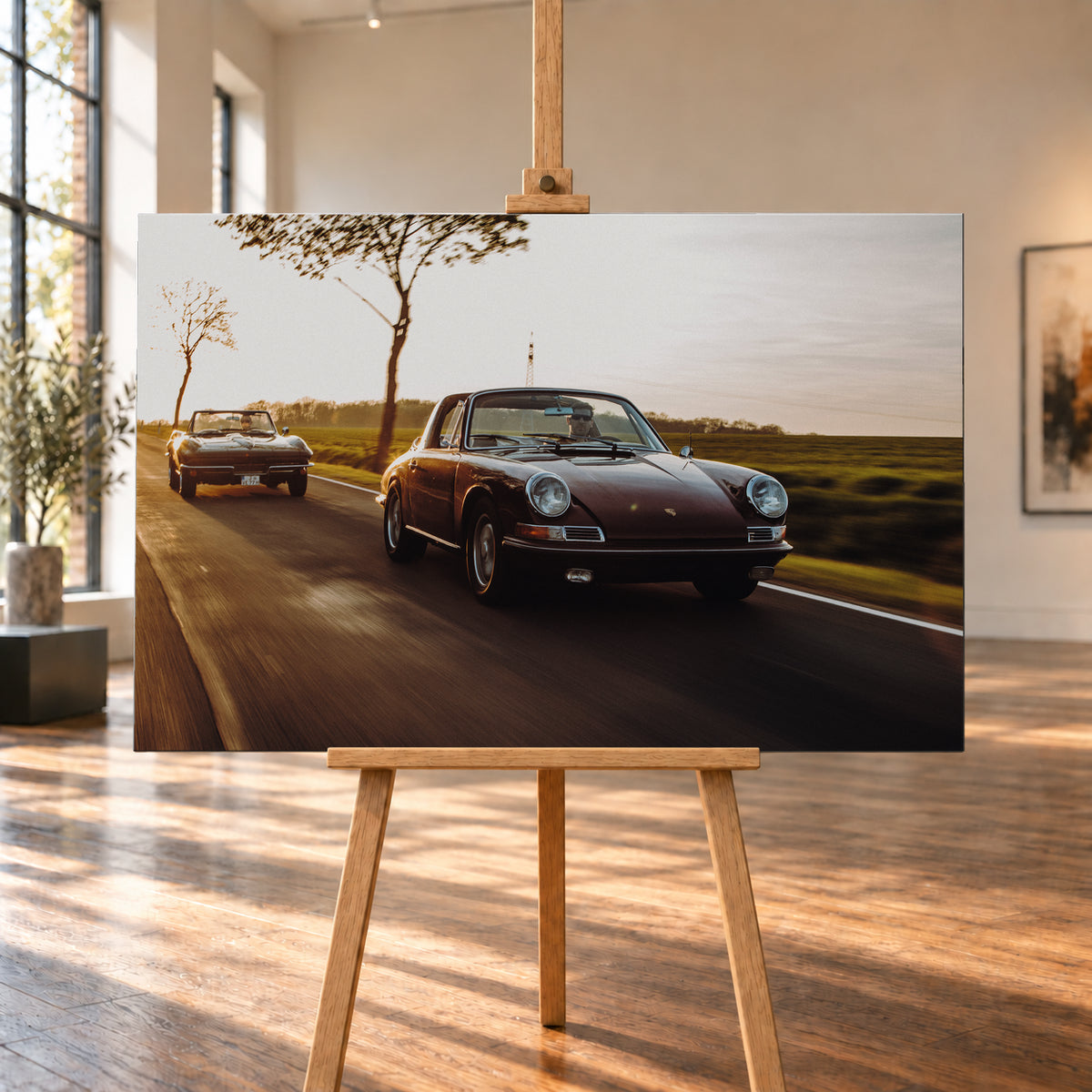 Ein dunkelrot wirkender Porsche 911 S Softwindow fährt rechts im Vordergrund, dahinter links eine dunkle Chevrolet Corvette C2; beide mit offenem Dach und sichtbaren Fahrern auf einer Landstraße bei tiefem, warmem Gegenlicht; Visualisierung als Leinwand-Wandbild, auf einer Holzstaffelei in einem hellen Galerieraum.