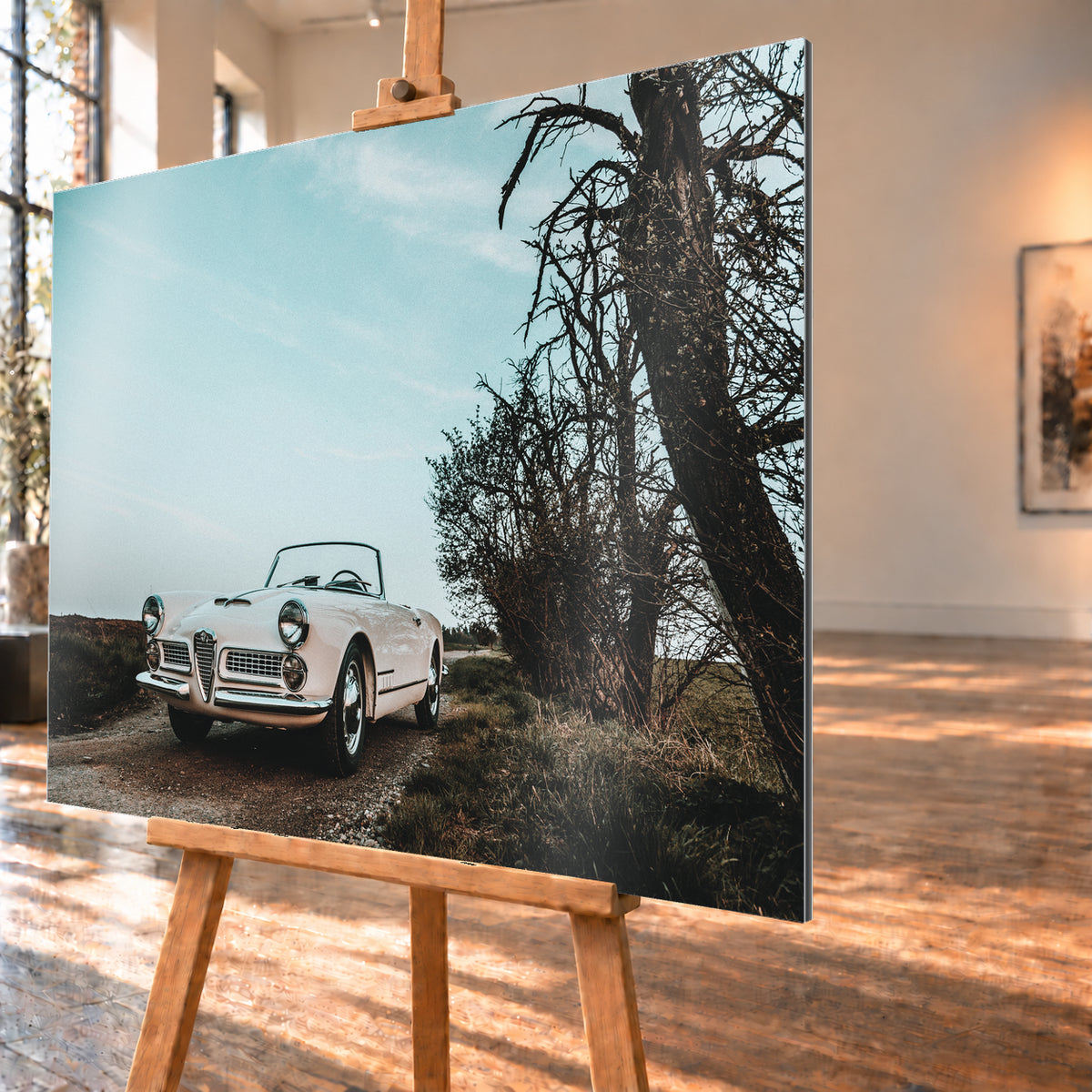 Heller Alfa Romeo 2000 Touring Spider in tiefer Front-Dreiviertelansicht auf einem schmalen Weg neben einem großen knorrigen Baum unter bläulichem, bewölktem Himmel; Visualisierung als Alu gebürstet-Wandbild, schräg von der Seite auf einer Holzstaffelei.