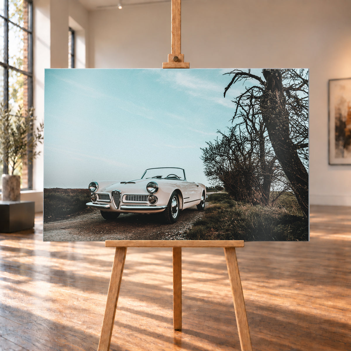 Heller Alfa Romeo 2000 Touring Spider in tiefer Front-Dreiviertelansicht auf einem schmalen Weg neben einem großen knorrigen Baum unter bläulichem, bewölktem Himmel; Visualisierung als Alu gebürstet-Wandbild, auf einer Holzstaffelei in einem hellen Galerieraum.