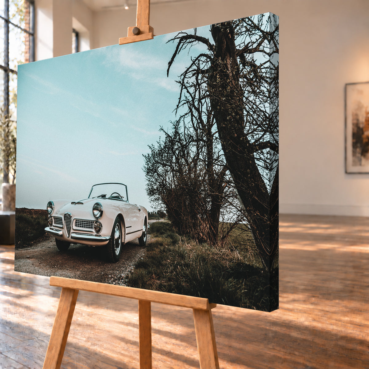 Heller Alfa Romeo 2000 Touring Spider in tiefer Front-Dreiviertelansicht auf einem schmalen Weg neben einem großen knorrigen Baum unter bläulichem, bewölktem Himmel; Visualisierung als Leinwand-Wandbild, schräg von der Seite auf einer Holzstaffelei.