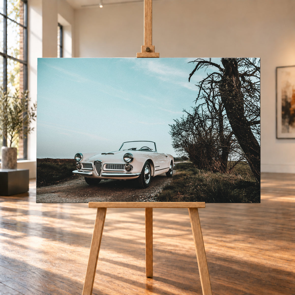 Heller Alfa Romeo 2000 Touring Spider in tiefer Front-Dreiviertelansicht auf einem schmalen Weg neben einem großen knorrigen Baum unter bläulichem, bewölktem Himmel; Visualisierung als Leinwand-Wandbild, auf einer Holzstaffelei in einem hellen Galerieraum.