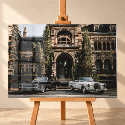 Dunkler 1964 Lincoln Continental links und heller 1965 Mercedes 300 SE W112 rechts, beide als offene Cabriolets in schräger Frontansicht vor einer ornamentierten historischen Steinfassade mit Rundbögen, hohen Fenstern und schlanken Bäumen; Visualisierung als Acrylglas-Wandbild, frontal auf einer Holzstaffelei.