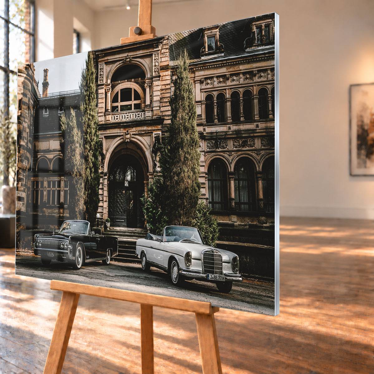 Dunkler 1964 Lincoln Continental links und heller 1965 Mercedes 300 SE W112 rechts, beide als offene Cabriolets in schräger Frontansicht vor einer ornamentierten historischen Steinfassade mit Rundbögen, hohen Fenstern und schlanken Bäumen; Visualisierung als Acrylglas-Wandbild, schräg von der Seite auf einer Holzstaffelei.