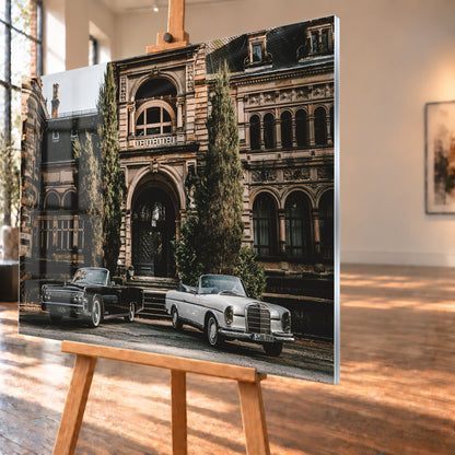 Dunkler 1964 Lincoln Continental links und heller 1965 Mercedes 300 SE W112 rechts, beide als offene Cabriolets in schräger Frontansicht vor einer ornamentierten historischen Steinfassade mit Rundbögen, hohen Fenstern und schlanken Bäumen; Visualisierung als Acrylglas-Wandbild, schräg von der Seite auf einer Holzstaffelei.