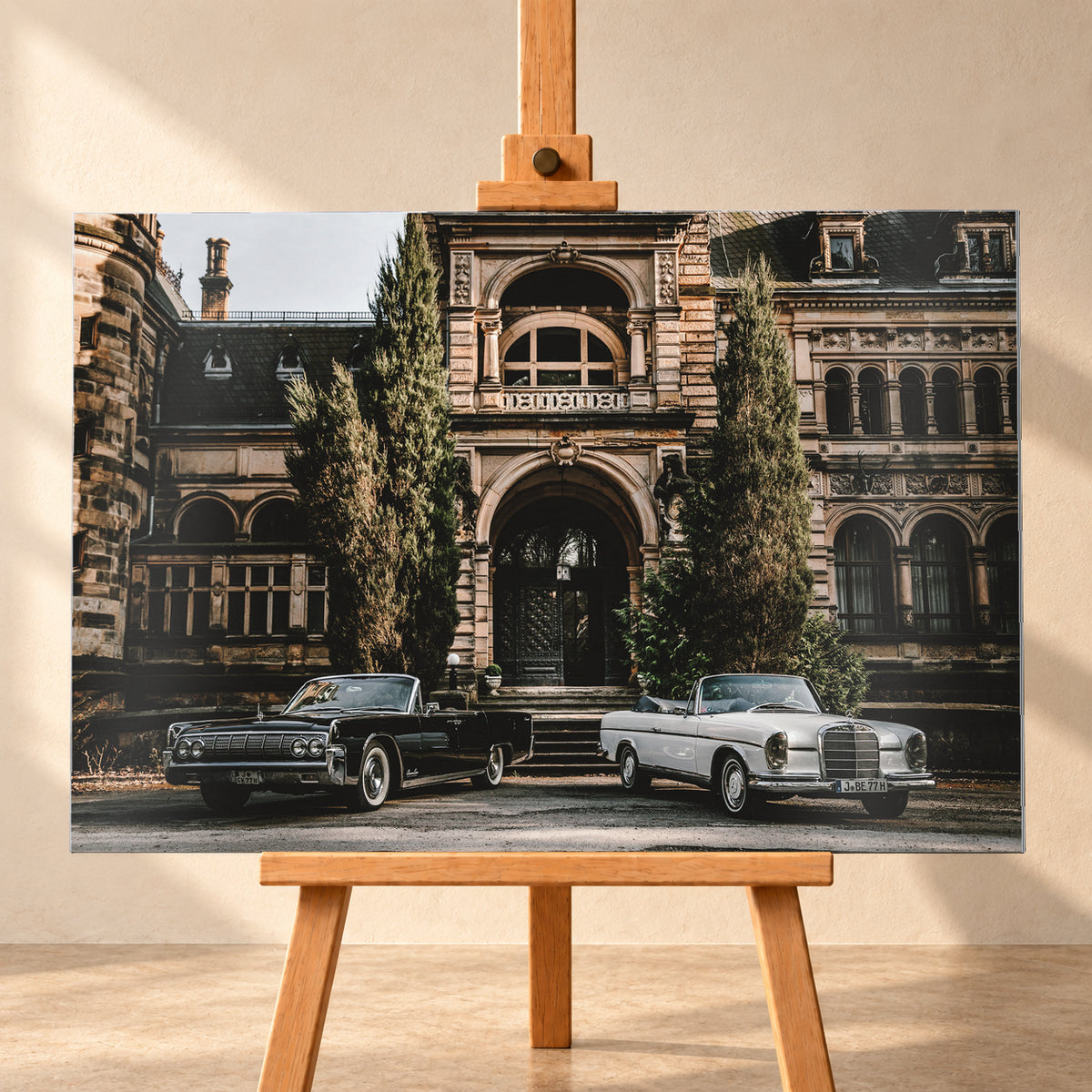 Dunkler 1964 Lincoln Continental links und heller 1965 Mercedes 300 SE W112 rechts, beide als offene Cabriolets in schräger Frontansicht vor einer ornamentierten historischen Steinfassade mit Rundbögen, hohen Fenstern und schlanken Bäumen; Visualisierung als Alu gebürstet-Wandbild, frontal auf einer Holzstaffelei.