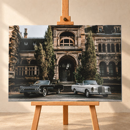Dunkler 1964 Lincoln Continental links und heller 1965 Mercedes 300 SE W112 rechts, beide als offene Cabriolets in schräger Frontansicht vor einer ornamentierten historischen Steinfassade mit Rundbögen, hohen Fenstern und schlanken Bäumen; Visualisierung als Alu gebürstet-Wandbild, frontal auf einer Holzstaffelei.