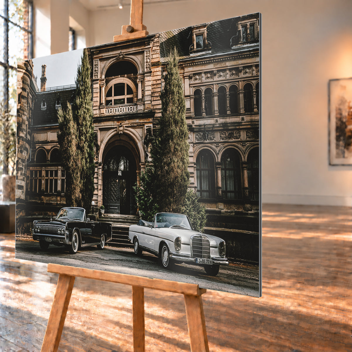 Dunkler 1964 Lincoln Continental links und heller 1965 Mercedes 300 SE W112 rechts, beide als offene Cabriolets in schräger Frontansicht vor einer ornamentierten historischen Steinfassade mit Rundbögen, hohen Fenstern und schlanken Bäumen; Visualisierung als Alu gebürstet-Wandbild, schräg von der Seite auf einer Holzstaffelei.