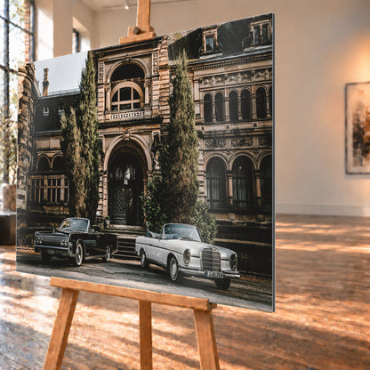 Dunkler 1964 Lincoln Continental links und heller 1965 Mercedes 300 SE W112 rechts, beide als offene Cabriolets in schräger Frontansicht vor einer ornamentierten historischen Steinfassade mit Rundbögen, hohen Fenstern und schlanken Bäumen; Visualisierung als Alu gebürstet-Wandbild, schräg von der Seite auf einer Holzstaffelei.