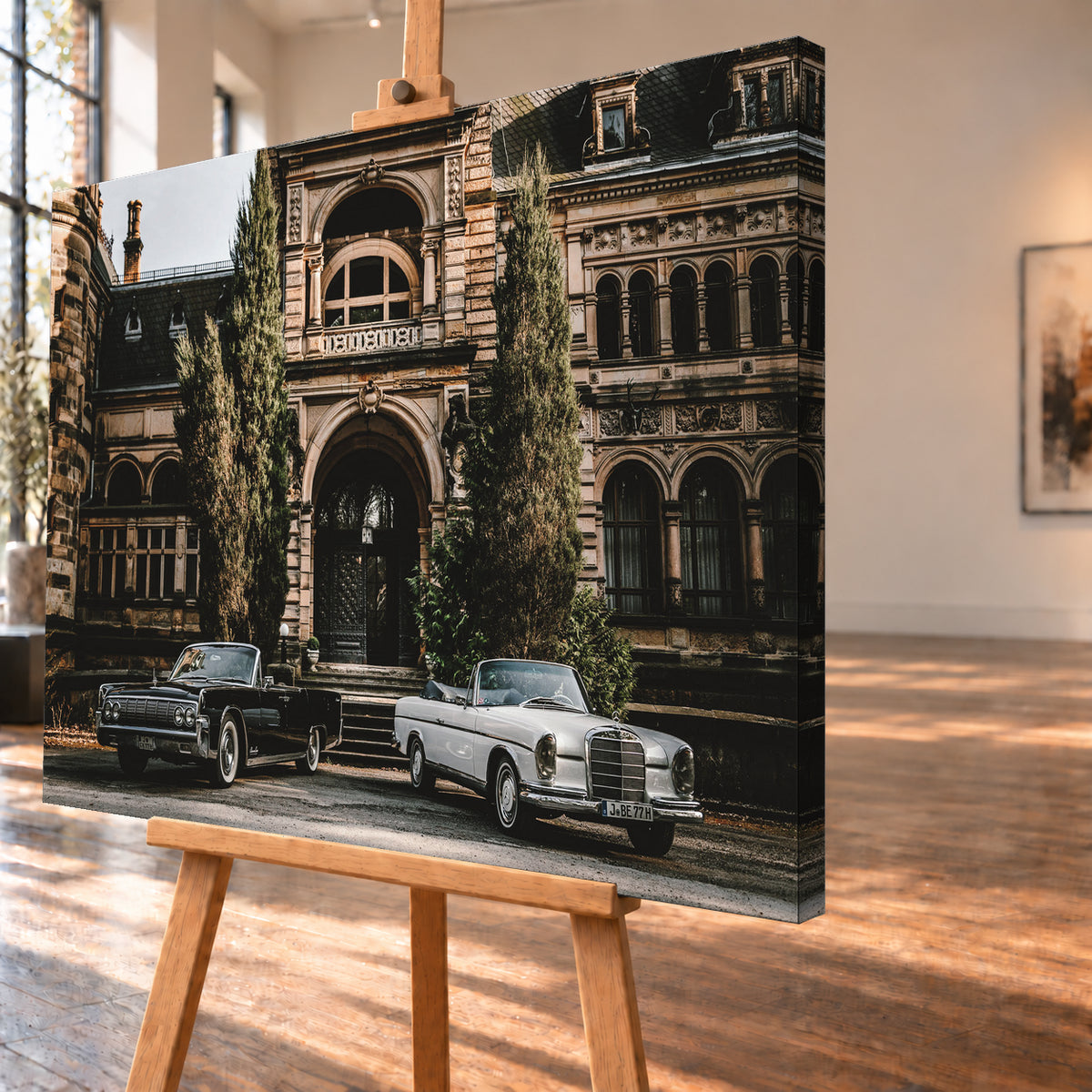 Dunkler 1964 Lincoln Continental links und heller 1965 Mercedes 300 SE W112 rechts, beide als offene Cabriolets in schräger Frontansicht vor einer ornamentierten historischen Steinfassade mit Rundbögen, hohen Fenstern und schlanken Bäumen; Visualisierung als Leinwand-Wandbild, schräg von der Seite auf einer Holzstaffelei.