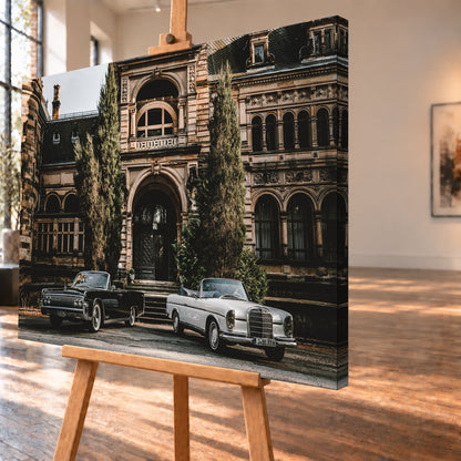 Dunkler 1964 Lincoln Continental links und heller 1965 Mercedes 300 SE W112 rechts, beide als offene Cabriolets in schräger Frontansicht vor einer ornamentierten historischen Steinfassade mit Rundbögen, hohen Fenstern und schlanken Bäumen; Visualisierung als Leinwand-Wandbild, schräg von der Seite auf einer Holzstaffelei.