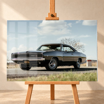 Dunkler Dodge Charger R/T in tiefer Dreiviertelansicht von vorn auf einer Straße unter hellem, wolkigem Himmel; im Hintergrund kahle Bäume und trockenes Gras; Visualisierung als Acrylglas-Wandbild, frontal auf einer Holzstaffelei.