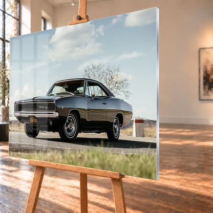 Dunkler Dodge Charger R/T in tiefer Dreiviertelansicht von vorn auf einer Straße unter hellem, wolkigem Himmel; im Hintergrund kahle Bäume und trockenes Gras; Visualisierung als Acrylglas-Wandbild, schräg von der Seite auf einer Holzstaffelei.