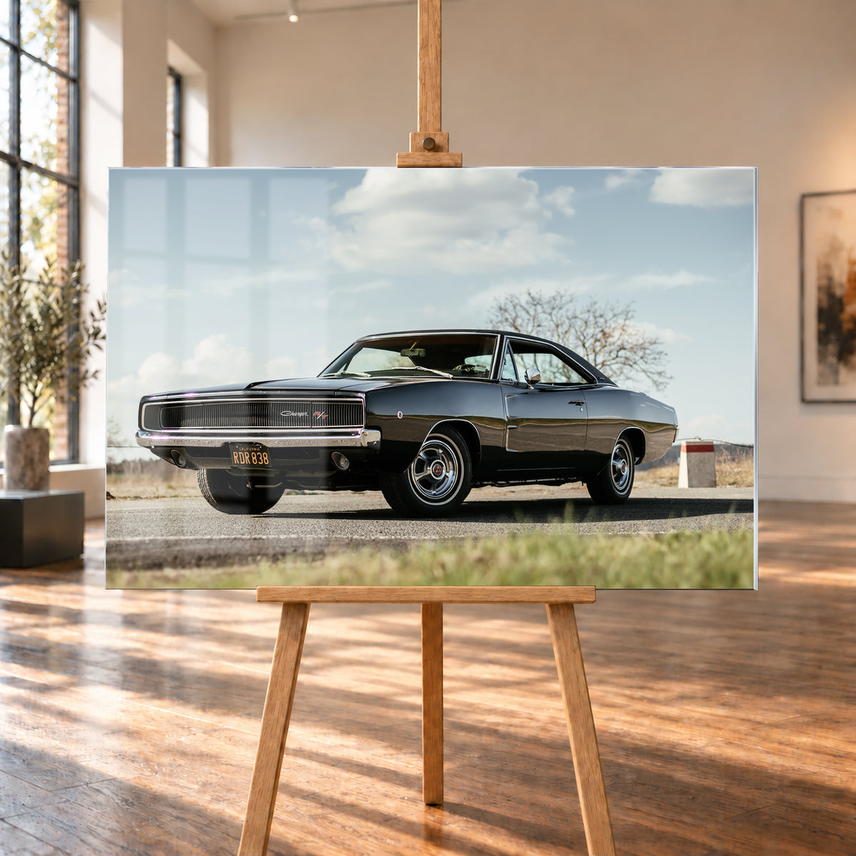 Dunkler Dodge Charger R/T in tiefer Dreiviertelansicht von vorn auf einer Straße unter hellem, wolkigem Himmel; im Hintergrund kahle Bäume und trockenes Gras; Visualisierung als Acrylglas-Wandbild, auf einer Holzstaffelei in einem hellen Galerieraum.