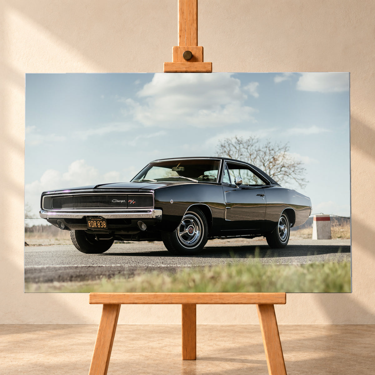 Dunkler Dodge Charger R/T in tiefer Dreiviertelansicht von vorn auf einer Straße unter hellem, wolkigem Himmel; im Hintergrund kahle Bäume und trockenes Gras; Visualisierung als Alu gebürstet-Wandbild, frontal auf einer Holzstaffelei.