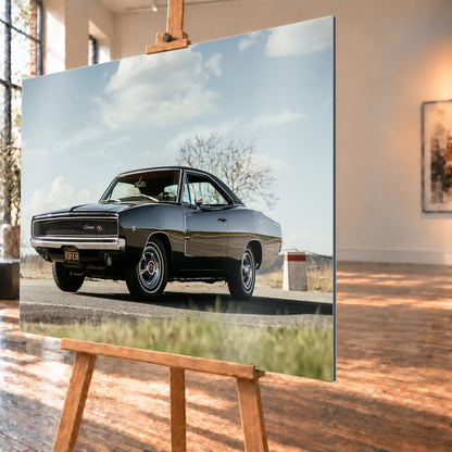 Dunkler Dodge Charger R/T in tiefer Dreiviertelansicht von vorn auf einer Straße unter hellem, wolkigem Himmel; im Hintergrund kahle Bäume und trockenes Gras; Visualisierung als Alu gebürstet-Wandbild, schräg von der Seite auf einer Holzstaffelei.