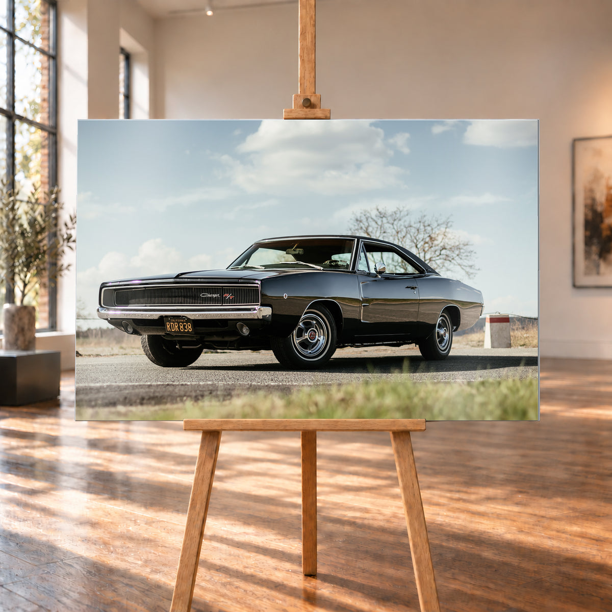 Dunkler Dodge Charger R/T in tiefer Dreiviertelansicht von vorn auf einer Straße unter hellem, wolkigem Himmel; im Hintergrund kahle Bäume und trockenes Gras; Visualisierung als Alu gebürstet-Wandbild, auf einer Holzstaffelei in einem hellen Galerieraum.