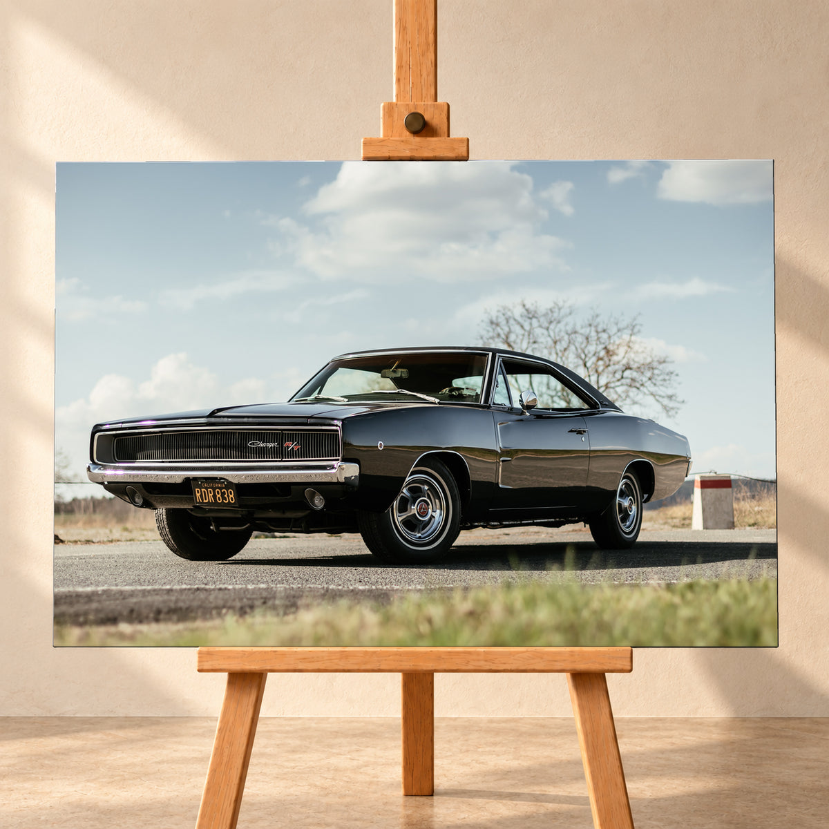 Dunkler Dodge Charger R/T in tiefer Dreiviertelansicht von vorn auf einer Straße unter hellem, wolkigem Himmel; im Hintergrund kahle Bäume und trockenes Gras; Visualisierung als Leinwand-Wandbild, frontal auf einer Holzstaffelei.