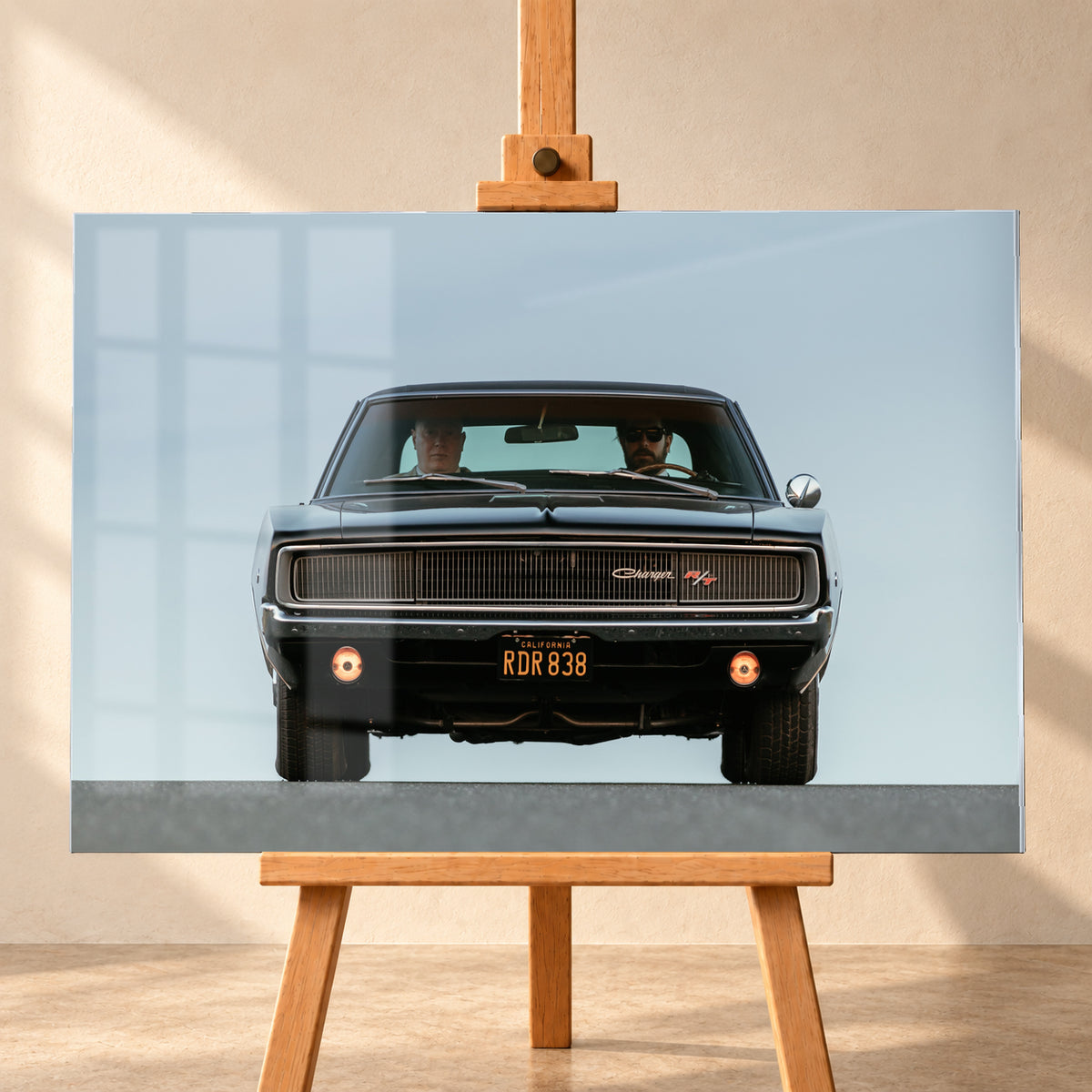 Sehr dunkler Dodge Charger R/T in tiefer, zentraler Frontalansicht vor blassblauem Himmel; zwei Personen sitzen hinter der Windschutzscheibe, zwei Zusatzleuchten glimmen warm; Visualisierung als Acrylglas-Wandbild, frontal auf einer Holzstaffelei.