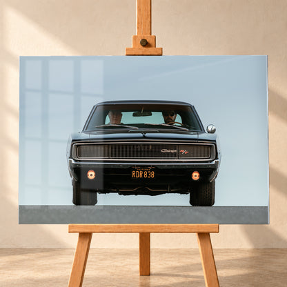 Sehr dunkler Dodge Charger R/T in tiefer, zentraler Frontalansicht vor blassblauem Himmel; zwei Personen sitzen hinter der Windschutzscheibe, zwei Zusatzleuchten glimmen warm; Visualisierung als Acrylglas-Wandbild, frontal auf einer Holzstaffelei.