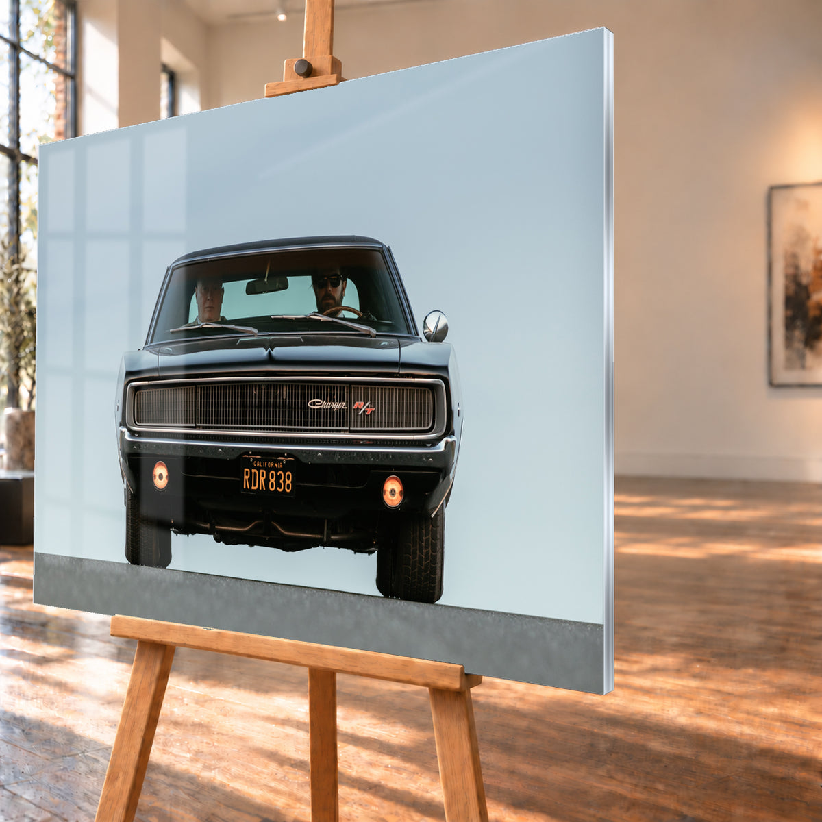 Sehr dunkler Dodge Charger R/T in tiefer, zentraler Frontalansicht vor blassblauem Himmel; zwei Personen sitzen hinter der Windschutzscheibe, zwei Zusatzleuchten glimmen warm; Visualisierung als Acrylglas-Wandbild, schräg von der Seite auf einer Holzstaffelei.